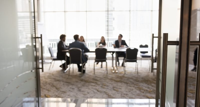 Group of businesspeople sitting at large conference table in negotiation room. Project partners brainstorming on ideas, discussing collaboration terms, teamwork. Business meeting blurred background